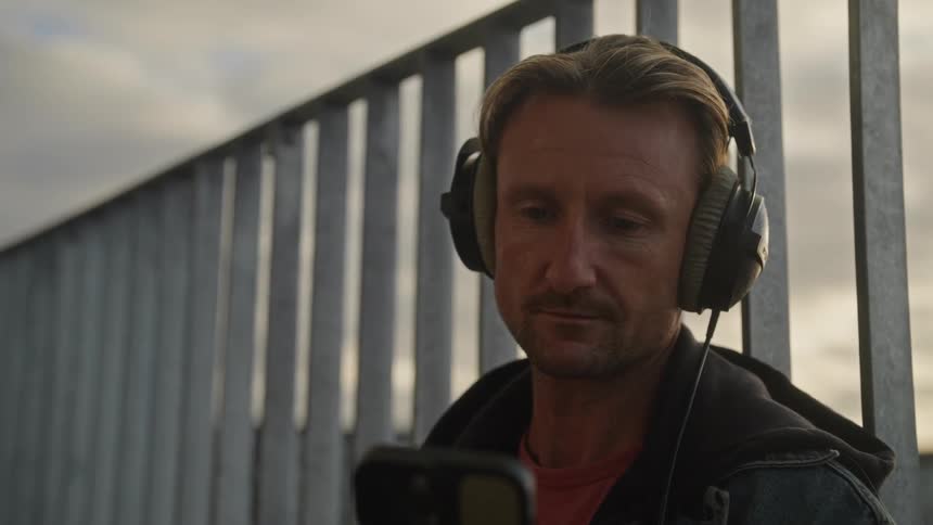 A man stands in front of a metal gate with headphones on holding a smartphone