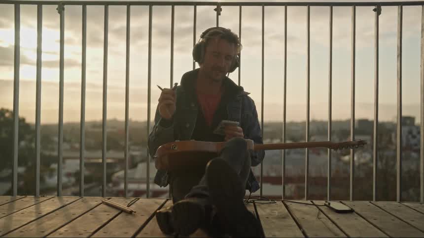 A man sitting on a balcony wearing headphones holding a guitar and listening to music