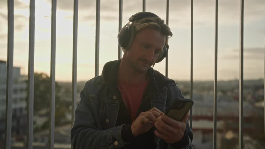 A man standing outside against a railing with headphones on while looking at his phone typing and smiling