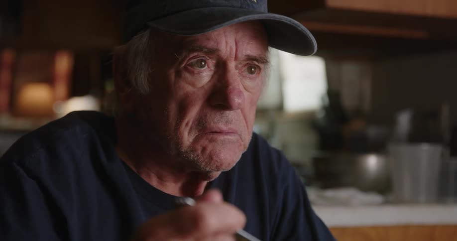 An old man wearing a cap reaches for a spoon then pauses and talks to somebody
