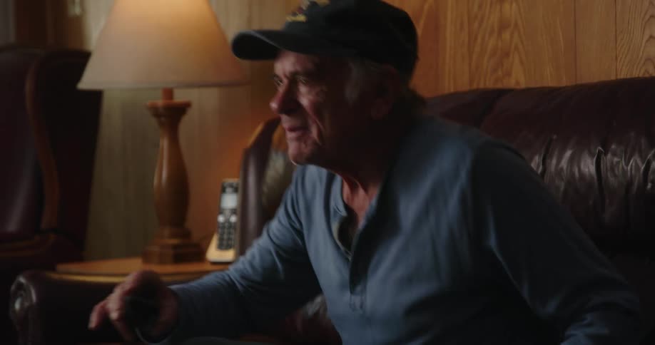 An older man sitting on his couch using the tv remote and sitting back while smiling