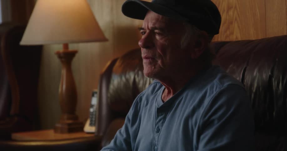 An older man sitting on his couch next to a lamp as he leans forward shouting angrily at the tv