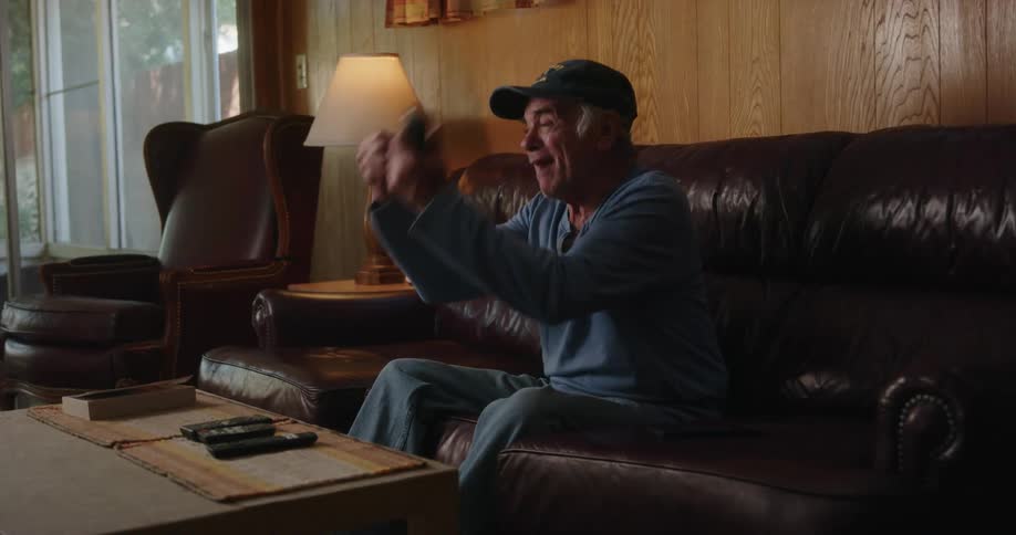 An older man sitting on his couch with a tv in his hand as he shouts and gestures aggressively