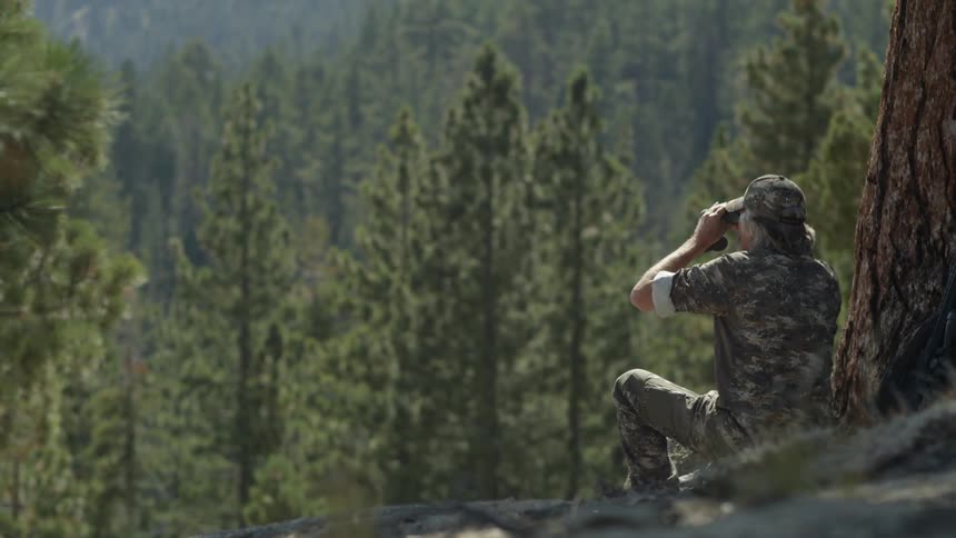 A man is sitting by a tree using binoculars