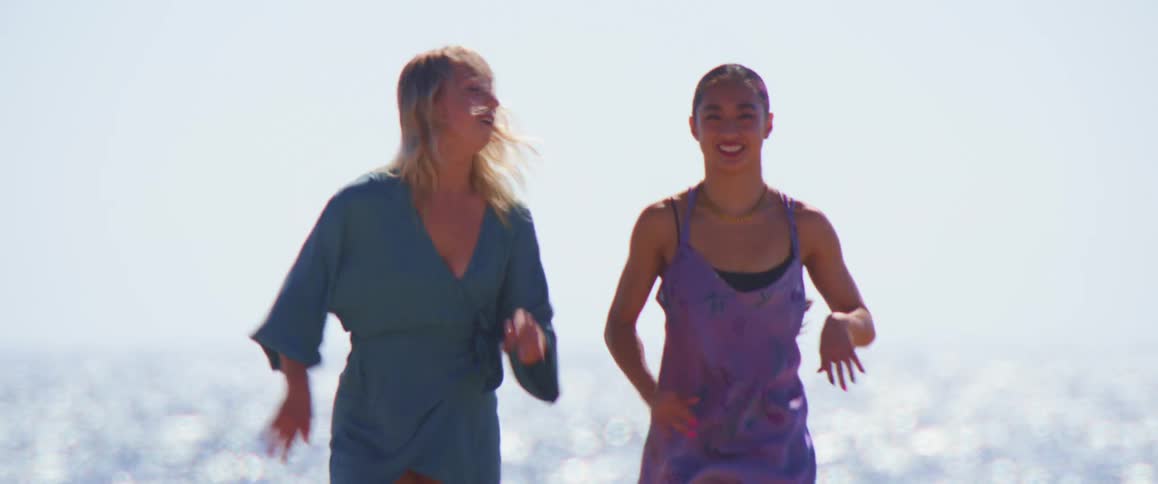 Two women in dresses run and laugh on a sandy beach