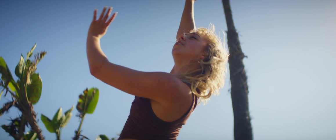 A woman dancing with arms above her head on a sunny day with palm trees behind her