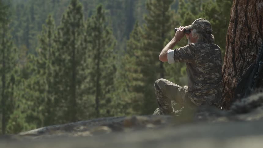 A man is sitting against a tree using binoculars
