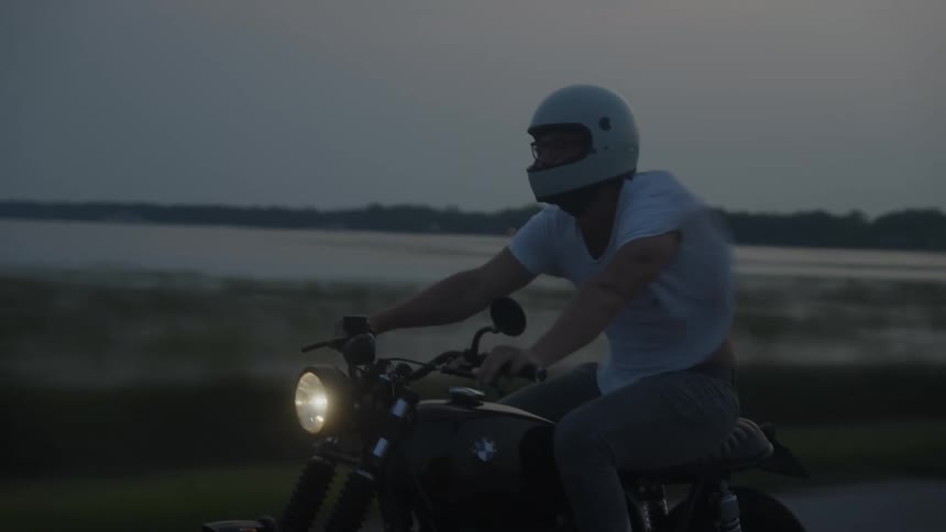 A man drives a motorcycle past a lake at dusk
