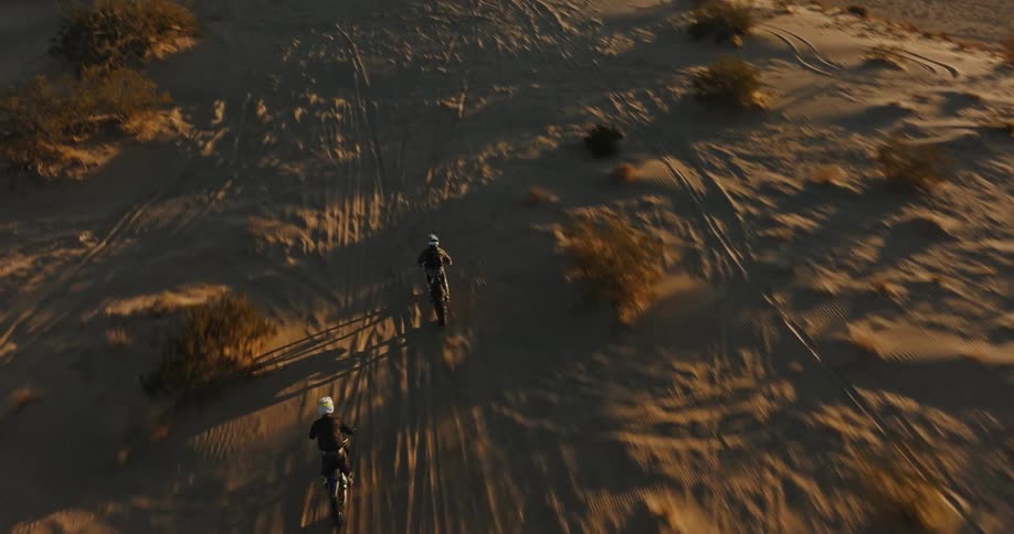 Two people riding motocross bikes across sand dunes at sunset