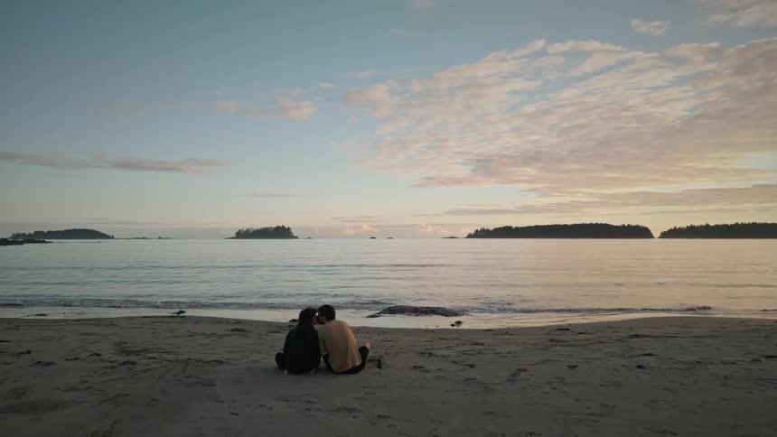 Young couple kiss while sitting in the sand beach at sunset next to the ocean