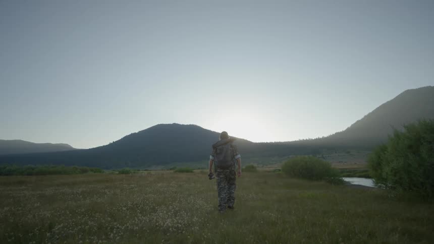 A hunter walking through a field next to a river at the foot of the mountains