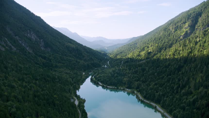 A beautiful view of treecovered mountain slopes on either side of an elongated lake of clear water reflecting the clouds in the sky overhead on a sunny day