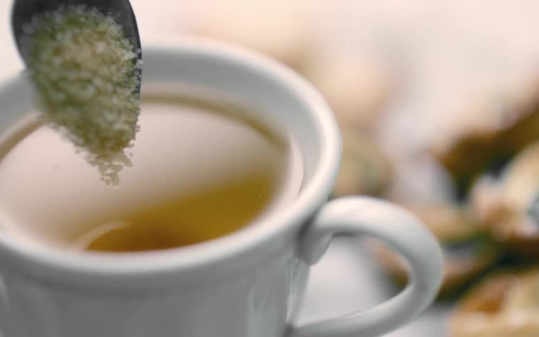 A spoonful of brown sugar crystals pour slowly in slow motion from a silver spoon into a white mug holding a brown liquid coffee or tea causing ripples as they enter the liquid