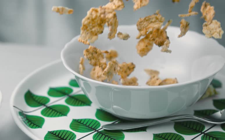 There is a white bowl of red strawberries and raspberries and the camera pans over to show bran flakes or corn flakes falling in slow motion into a cereal white bowl on top of a plate decorated with green leaves next to a spoon