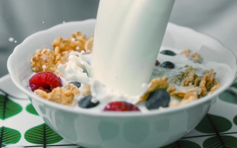 A stream of milk is pouring into a bowl of cereal with raspberries and blueberries