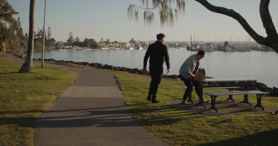 Male friends greet each other with a hug before sitting down at picnic table on shore together