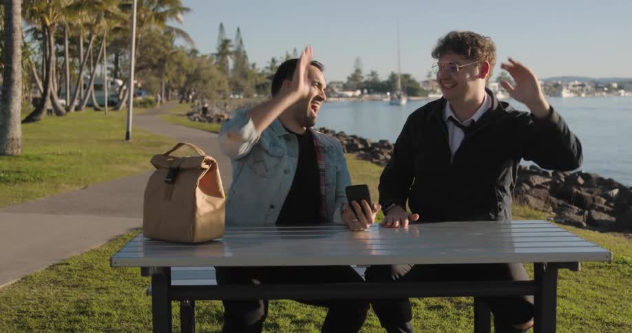 Men sitting at picnic table on shore high five while viewing smartphone screen