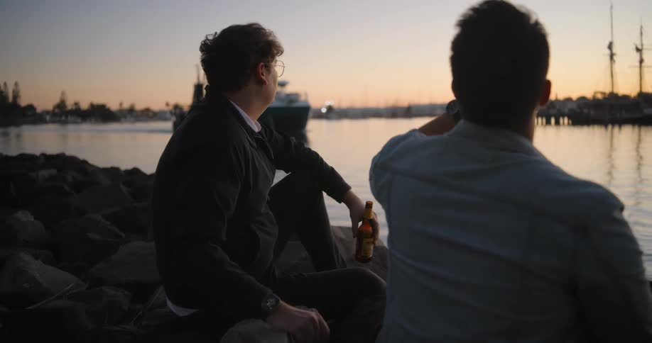 Two men sit on rocks at a lake drinking beer