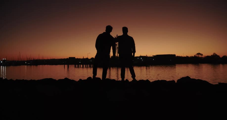 Two men stand next to a river and throw stones at sunset