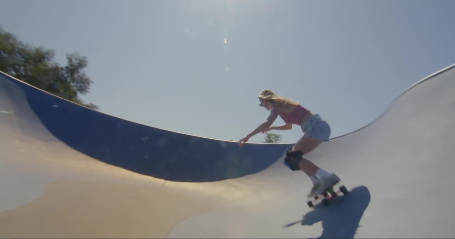 A young blonde woman is skateboarding along the concrete track at a skate park on a sunny day