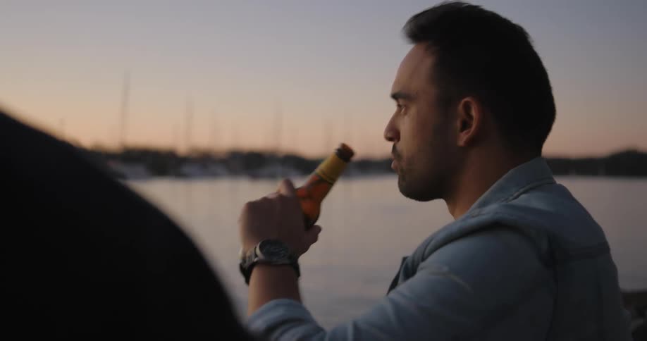 A man talking to another in front of a lake and then drinking a beer