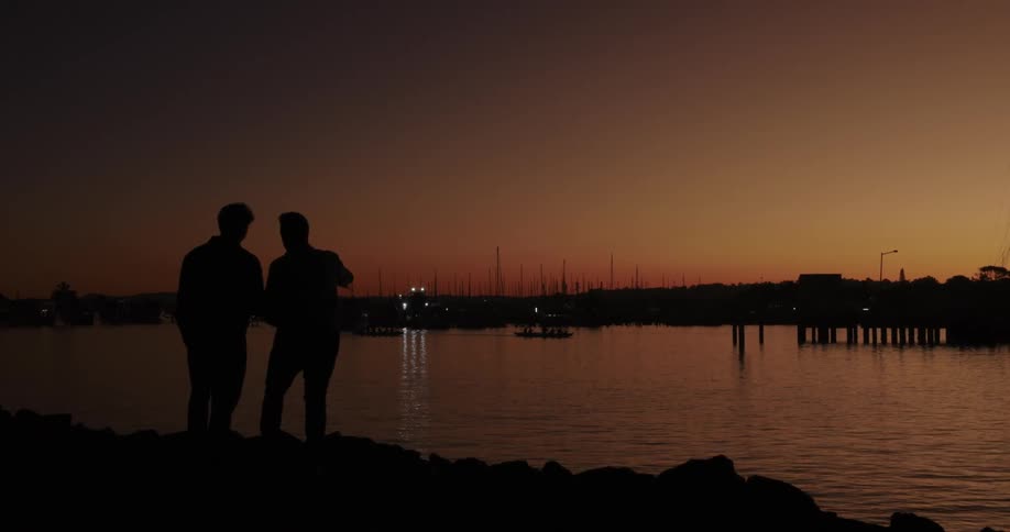 Silhouette of two friends by the river conversing under a reddish sky