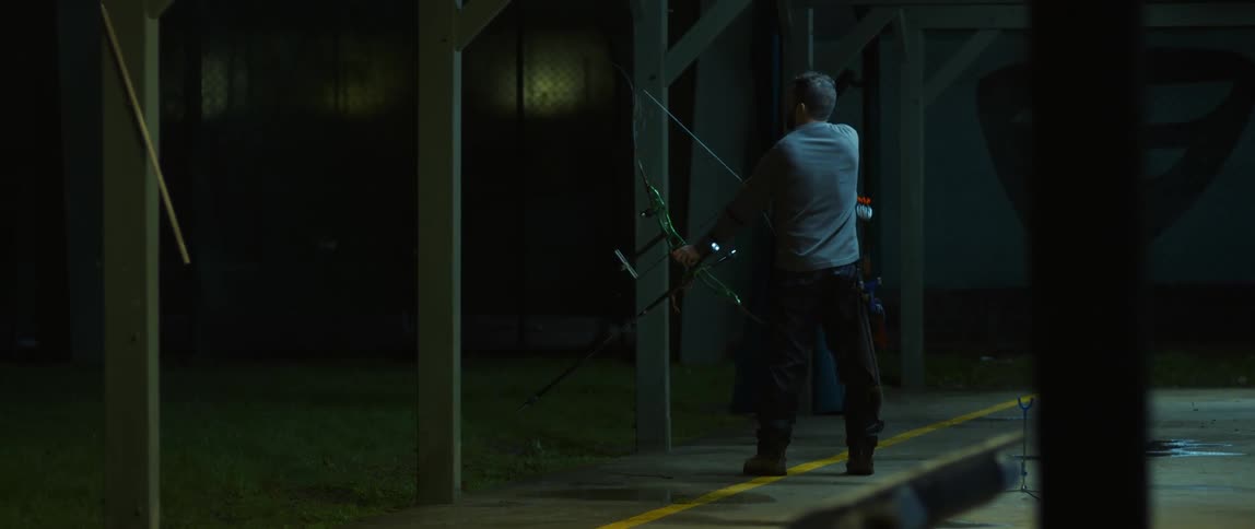 A bearded man wearing a gray blouse gets ready to shoot an arrow from a bow in a garage