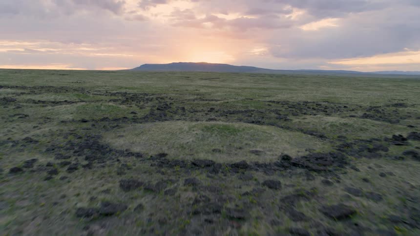 Skimming above a primordial landscape of honeycomb rock formations and rippled sagebrush mounds as the sun rises behind a mountain ridge in the distance