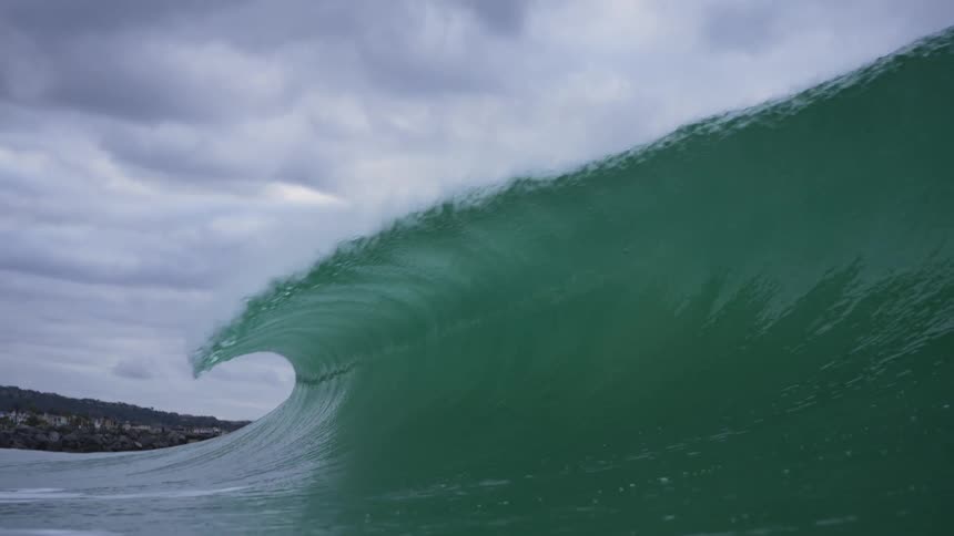 A huge waves splashes as it curls and rolls into shore under a cloudy sky and the view moves underwater