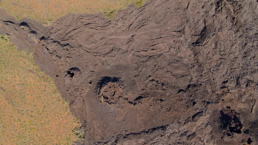 Birds eye view of craters and vents that line a lobe of basaltic lava extending into a colorful field of sage and yellow wildflowers