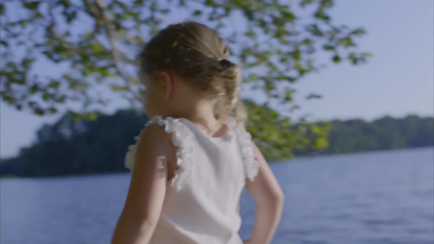 Little girl with a braid wearing a white top and skirt strolls by the lake