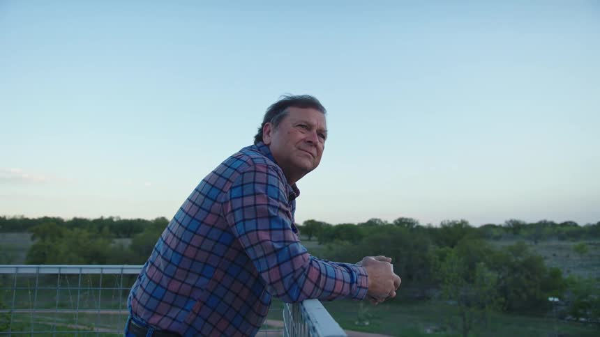 A man wearing a jean and a long sleeve standing close to a handrail made from mech steel in a balcony with vegetation and trees seen to the ground