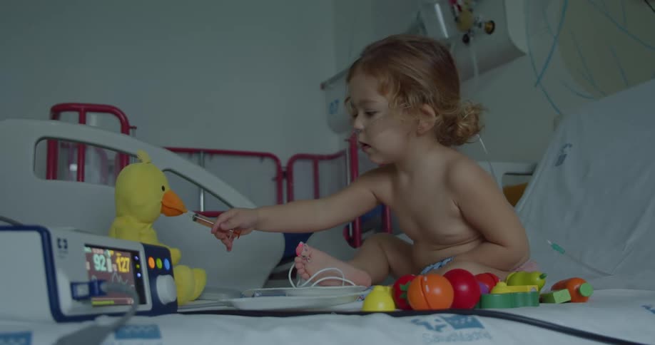 A very young girl plays with a stuffed animal and toys while in a hospital bed