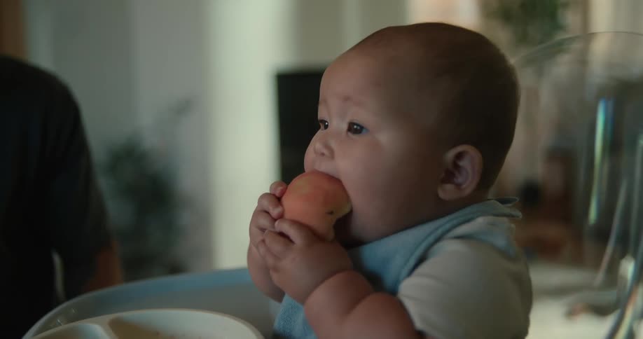 A baby sits eating a fruit while his parent looks at them smiling