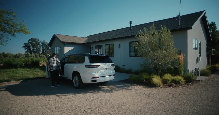 A white suv pulling up close to a home in the suburban with a white man and a woman stepping out as the man takes out a box from the trunk of the car