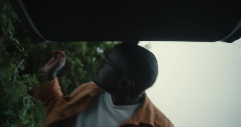 Man with a white top and a brown shirt places a bag in the trunk of the car