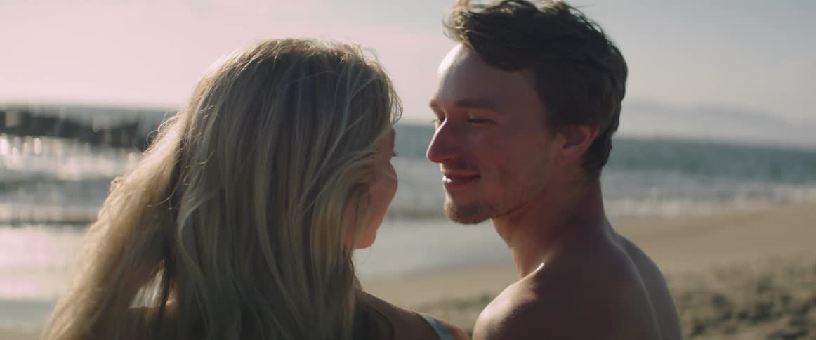 A man and a blonde woman kiss in front of the water on the beach
