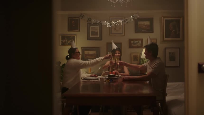 Three people wearing party hats indoors sitting at a table and toasting with champagne glasses for a birthday party during night time