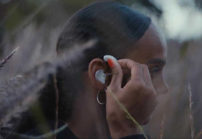 A woman sitting in a grassy field is putting a hearing aid in her ear
