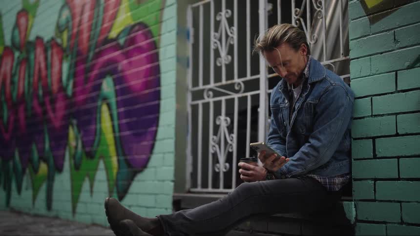 A blond man sits drinking from a glass and then typing on a cell phone