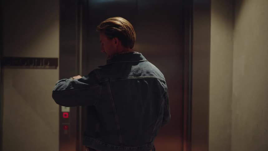 A blonde man in a denim jacket waits for the elevator and checks his watch