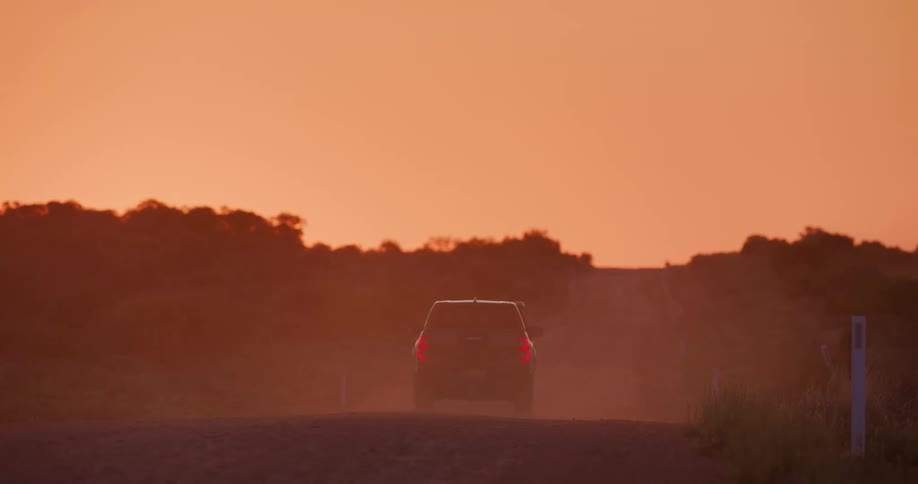 A black truck travels on a dirt road in the rain with an orange sunset sky in the distance