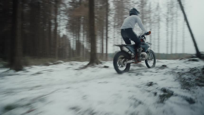 A cyclist rides a motorcycle through a snow covered forest and over a ramp leaping into the air