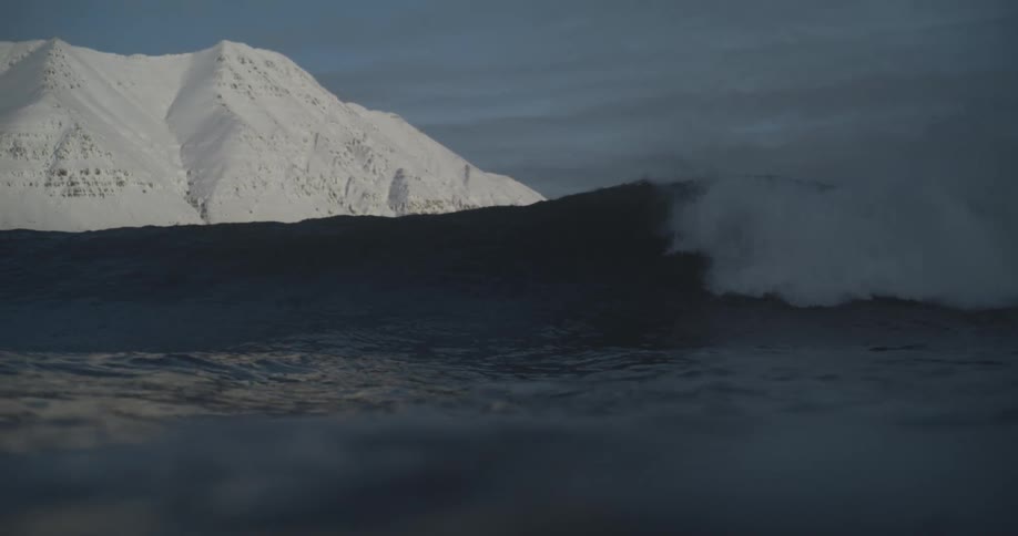 An ocean wave crashes against the shore its force contrasted by the serene backdrop of a snowcapped mountain