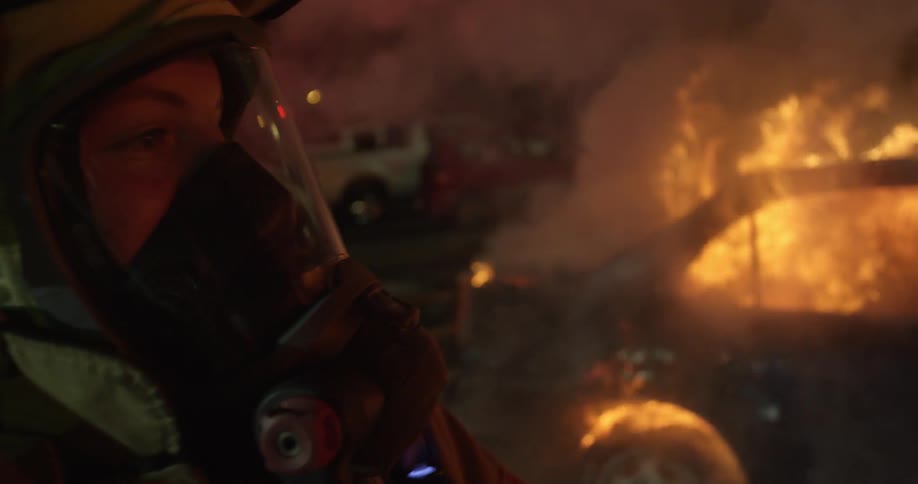 A firefighter holding a hose and dousing a raging car fire at night