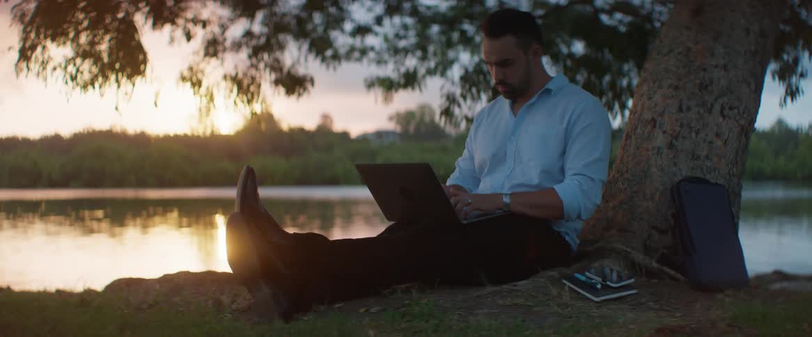 A man sitting on the grass typing on a laptop at sunset