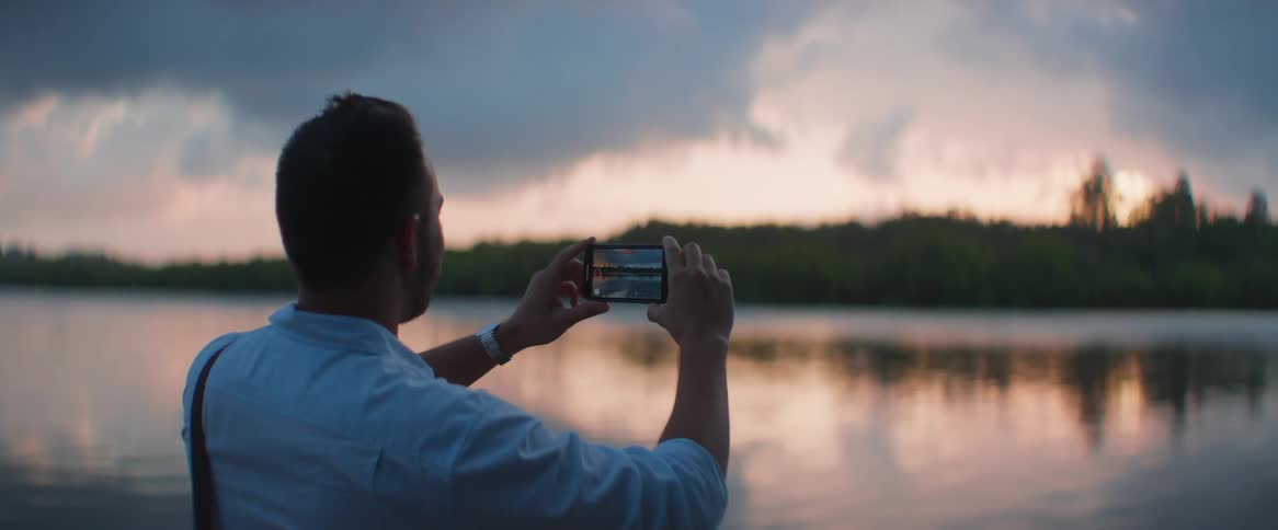 A man stands near a lake at dusk videotaping the surroundings with a mobile phone