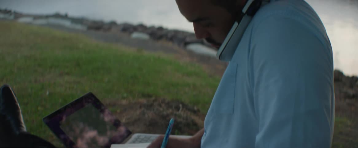 A man sitting on the grass with a laptop and writing in a notebook