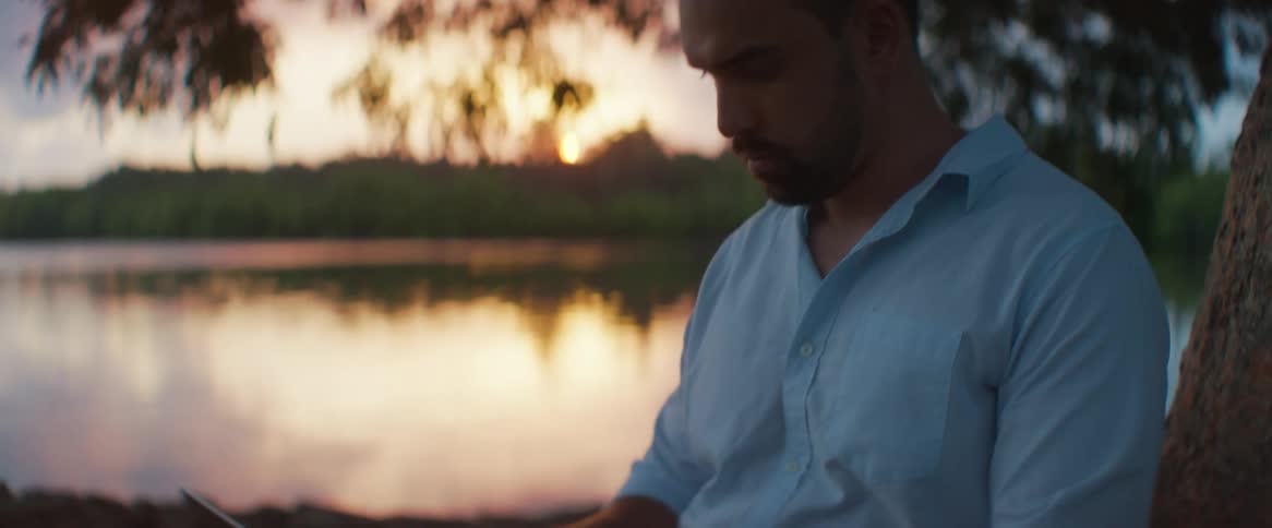 Man types on a laptop while sitting under a tree at dusk