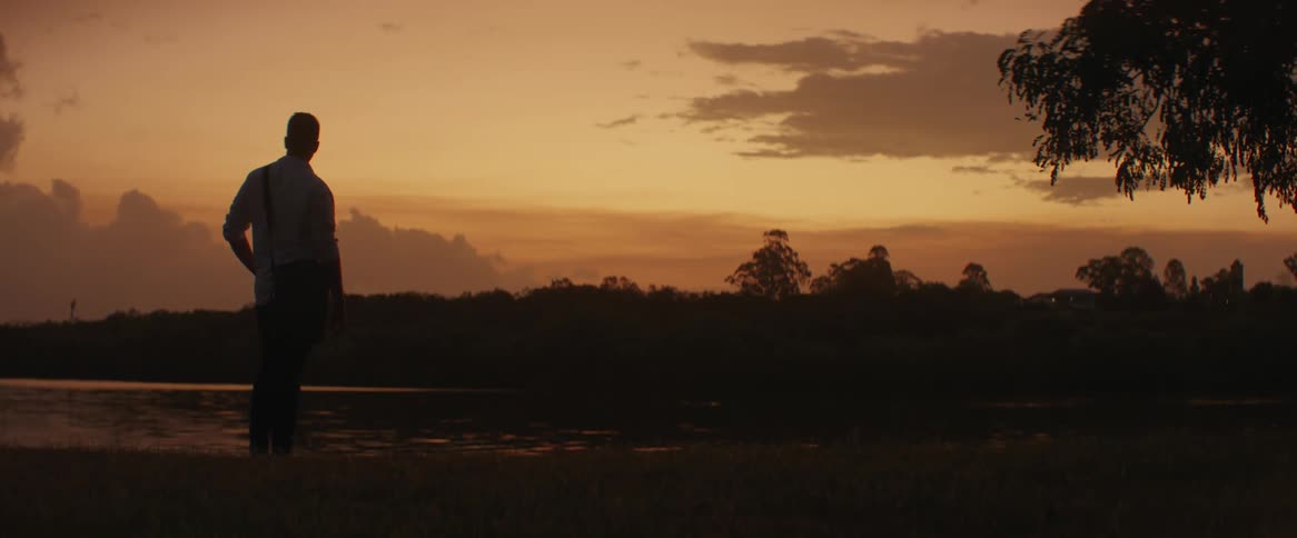 A man with a side bag stands near a river looking around at sunset